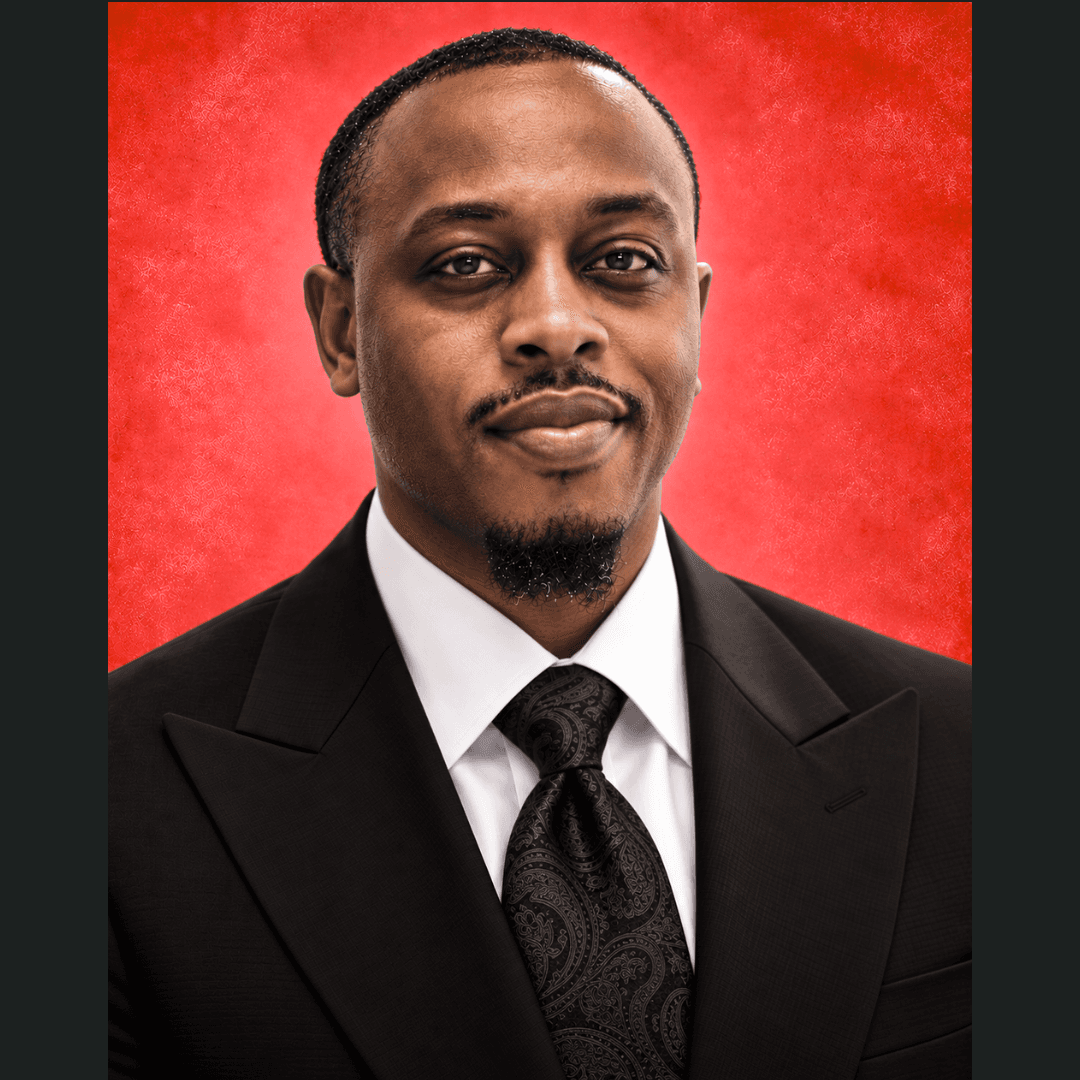 Black man with a goatee in a black suit and paisley tie, red background.
