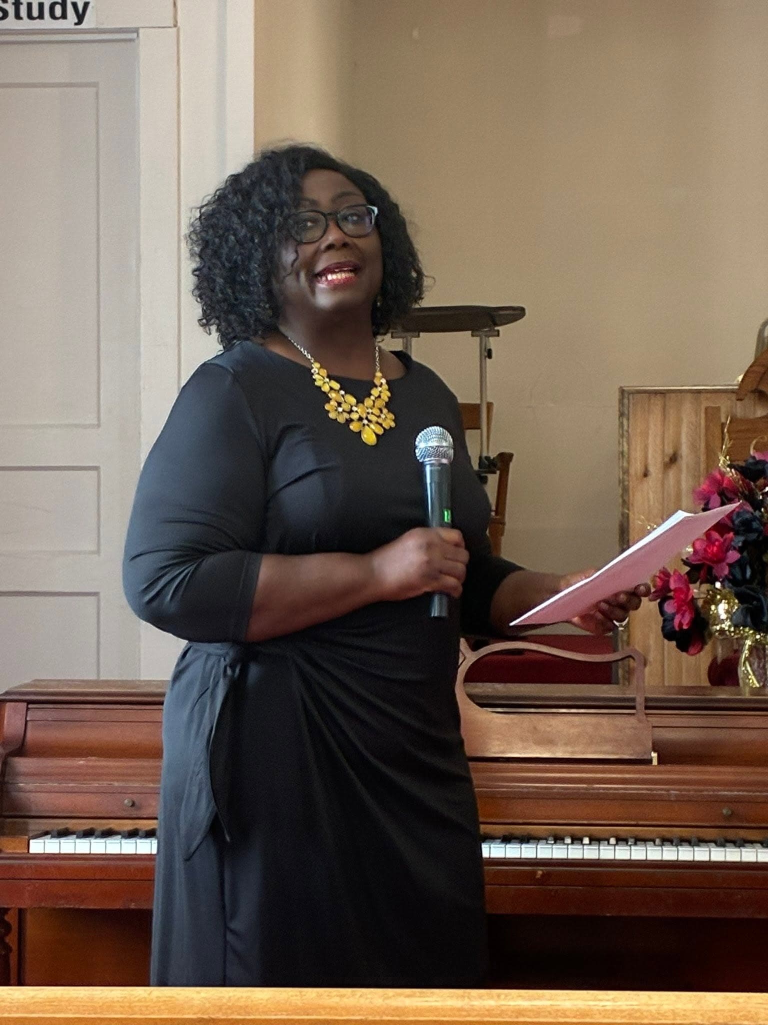 Smiling Black woman in black dress and yellow necklace holding microphone and paper near piano.