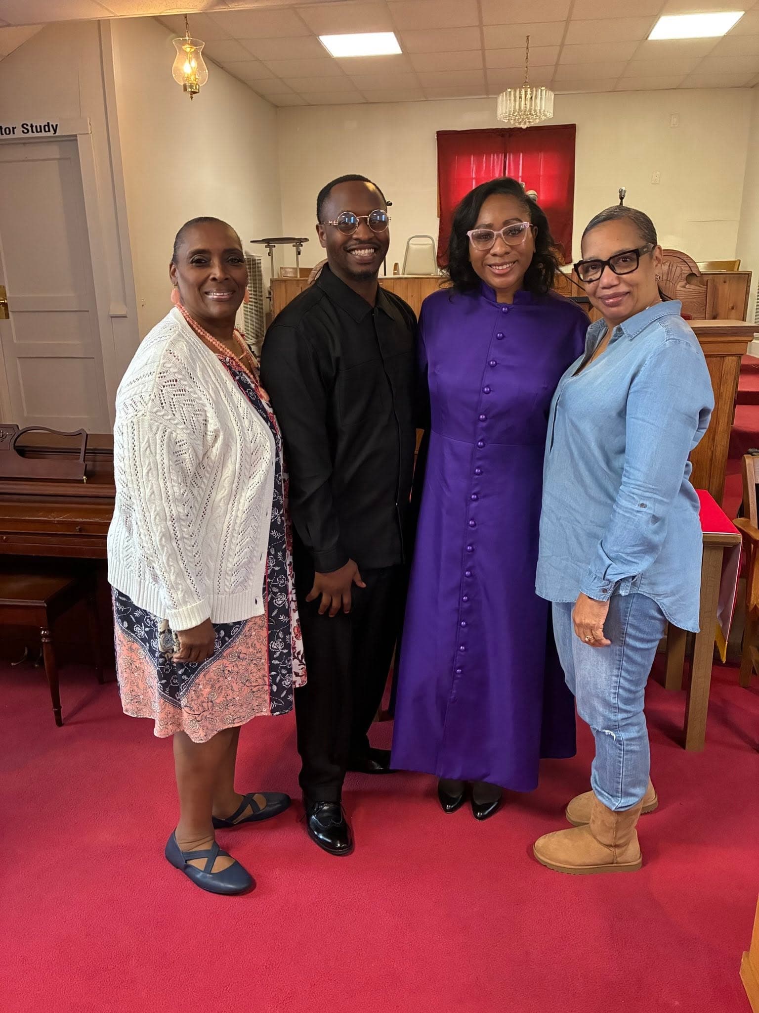 Four smiling people pose in a church, including a woman in a long purple robe.