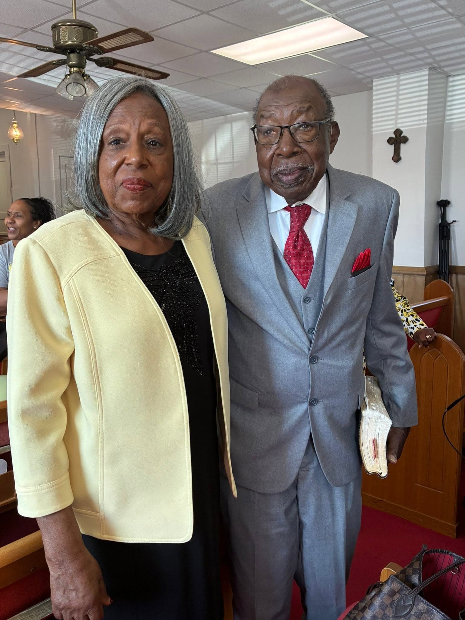 Elderly Black couple in church; man in grey suit holding Bible, woman in yellow jacket.