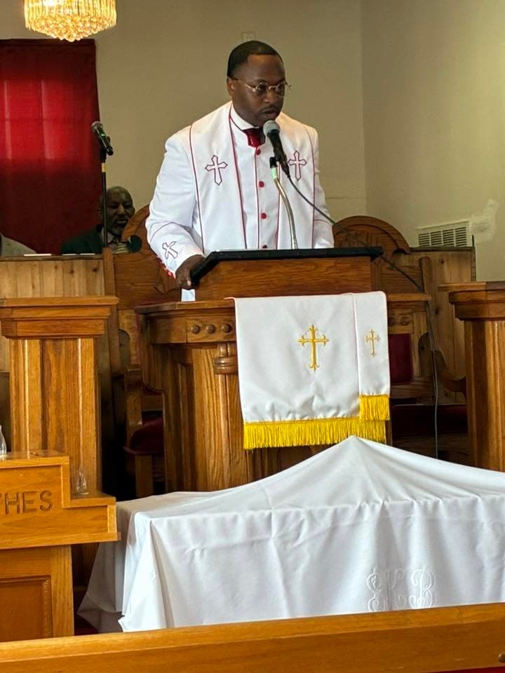 A Black minister in white clerical robes speaking into a microphone at a wooden pulpit.