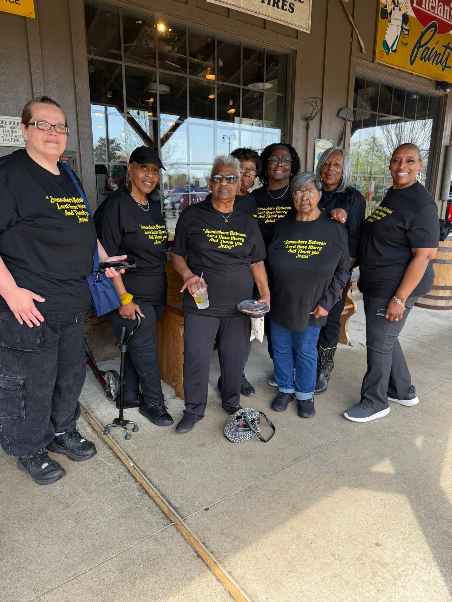 Seven women pose on a porch wearing matching black t-shirts with yellow religious text.